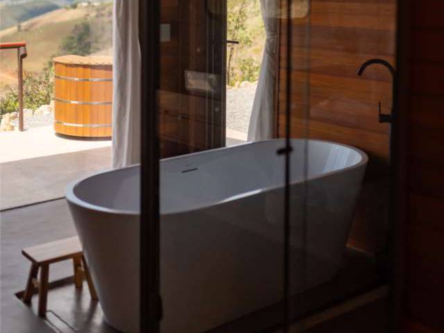 Banheira de imersão branca da Cabana Marama diante da janela, com o ofurô de madeira e o vale ao fundo