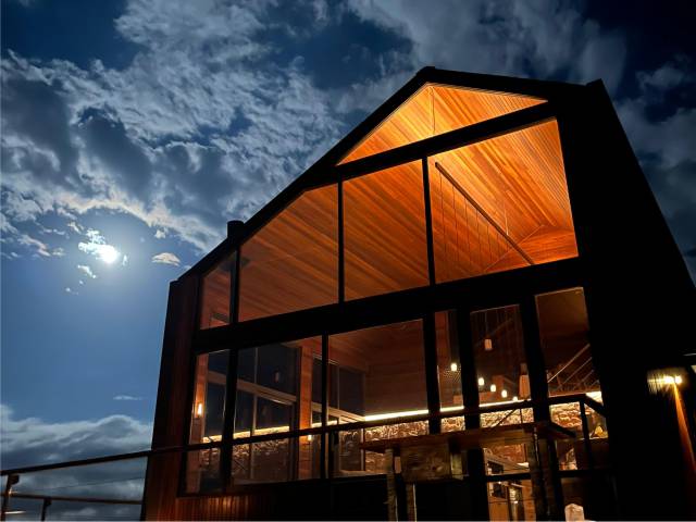 Fachada de vidro da Cabana Marama iluminada à noite, sob o luar e as nuvens